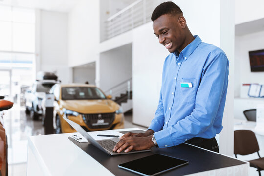 Positive black car salesman using laptop computer at work desk in auto dealership. Cheerful African American automobile manager working with pc in showroom store, free space
