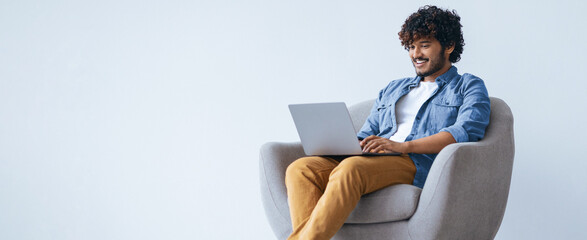 Naklejka premium A young man is sitting in a chair with a laptop on his lap. He is smiling while looking at the screen. This scene takes place in a bright and simple room during the day.