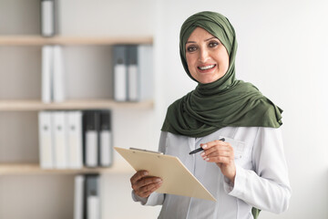Happy Muslim Female Doctor Posing Smiling To Camera Holding Medical Case Record Standing In Modern Clinic Office Indoors. Portrait Of Mature Middle-Eastern Professional Therapist Lady Wearing Hijab