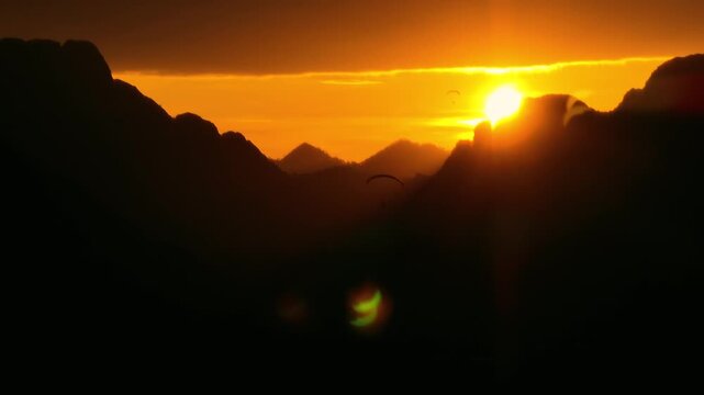 Paraglider flying above dramatic mountain landscape during golden hour at sunset. Scenic aerial view with warm sunlight creating a cinematic and adventurous atmosphere