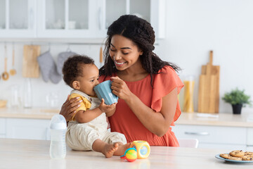 Fototapeta premium Caring Black Mom Teaching Cute Infant Baby To Drink From Cup While Spending Time Together In Kitchen At Home, Adorable Toddler Child Sitting On Counter Table And Enjoying Tasty Beverage, Closeup