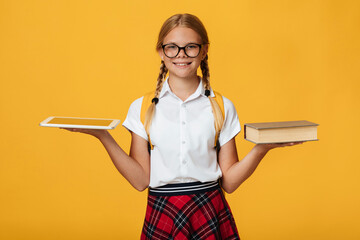 Cheerful young caucasian schoolgirl with pigtails, backpack in glasses hold book and tablet and chooses, isolated on yellow background. Modern education, digital devices, back to school and new normal