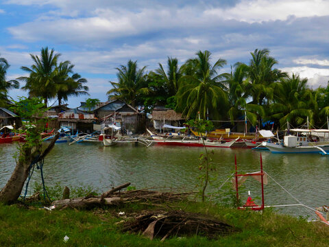 Fishing boats on the river. Traditional fishing boats moored along a tropical village shoreline. Colorful bancas rest on calm water beside palm trees, wooden houses, and a coastal community.