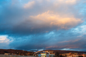a massive, textured cloud formation dominating the blue sky, with soft sunlight hitting its peak, hovering over a coastal town and distant brown hills, Bulgaria
