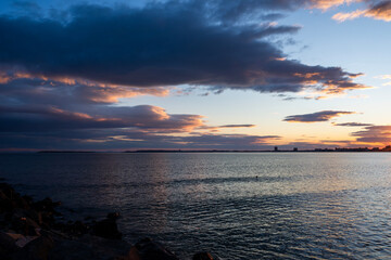 a calm body of water at twilight, with a dramatic sky of dark, layered clouds illuminated by touches of orange sunlight over a distant city skyline, Sunny beach, Bulgaria
