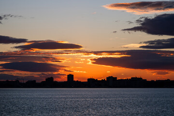 a dramatic sunset over a calm body of water, with dark, layered clouds glowing orange and yellow above a silhouetted city skyline.