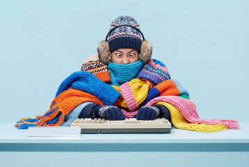 Funny woman freezing at work desk wrapped in many colorful wool scarves and hats typing on keyboard during heating failure on blue background.