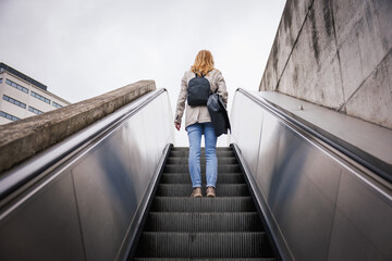Woman with laptop bag and backpack commuting to work on escalator in urban area. Daily commute and...