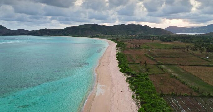 Lombok Pantai Tomang Omang beach stretching along a turquoise ocean with coastal mountains and fertile agricultural land, the island diverse tropical landscape under a cloudy sky. Aerial drone flight