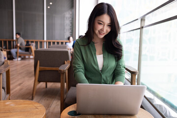 A smiling young Asian woman working on her laptop in a bright, modern cafe. She wears a green cardigan, reflecting a productive freelance lifestyle and remote work culture in a cozy atmosphere