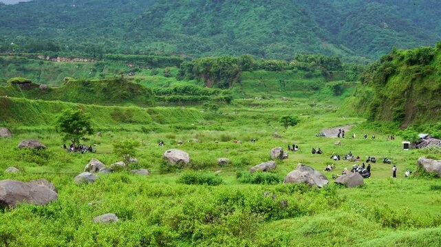 Scenic Ranu Manduro grassland landscape filled with large scattered boulders and Mount Penanggungan backdrop. Busy tourist spot with visitors relaxing on green meadow in Mojokerto, East Java.