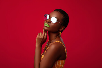 Sensual young afro american woman in summer outfit and stylish sunglasses posing on red studio background, closeup profile portrait, copy space. Eyeglasses, eyewear, spectacles concept © Prostock-studio