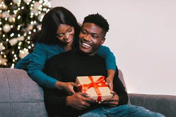 A young couple sits on a sofa smiling at each other as they share a special moment during the holidays. The woman hugs the man from behind while he holds a gift wrapped with a red ribbon.