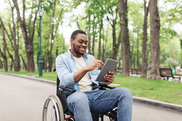 Obraz premium Happy young black man in wheelchair using tablet pc, browsing web or watching movie at spring park. Millennial African American guy checking social media on mobile device outdoors