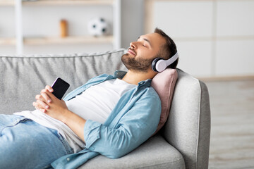 Relaxed young guy lying on couch in headphones, listening to music on smartphone, using new mobile app, mockup. Calm Caucasian man enjoying audio book or popular song at home