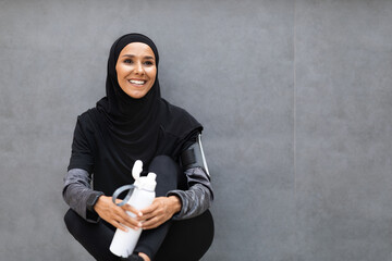 Happy young arab female athlete in hijab, sportswear with smartphone and bottle of water on gray wall background. Jogging in morning, tech for training, sports, body care during covid-19 pandemic