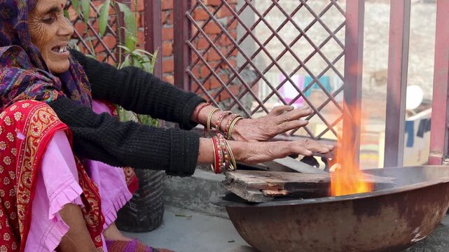 An Indian village woman warms her hands near an open fire, showing winter cold, traditional lifestyle, and everyday rural life practices in an Indian village setting.