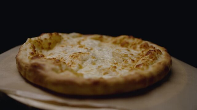 Traditional georgian cheese-filled bread, megruli khachapuri, rotating on a wooden board against a black background. Appetizing golden-brown crust