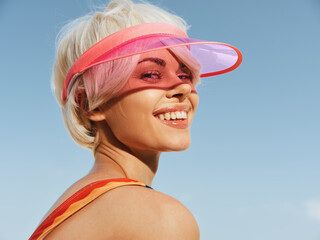Close portrait of a smiling woman wearing a pink translucent visor outdoors under a bright sky,...