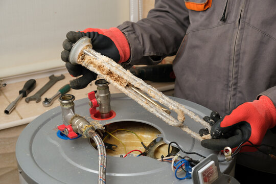 Repairman in red gloves demonstrates a burnt-out boiler heating element covered with lime coating. Electric Water Heater Maintenance and Repair.