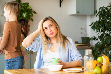 Young woman mom sitting on a kitchen table looking stressed suffers from headache because of noise...