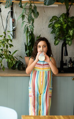 A young girl in a colorful striped jumpsuit drinks milk in a cozy kitchen filled with green...