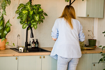 Woman washing plates dishes at a modern kitchen sink, surrounded by green indoor plants, wooden...