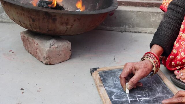 An elderly woman writes on a slate with chalk while a wood fire burns nearby for warmth, showing lifelong learning, resilience, and rural winter life in India.
