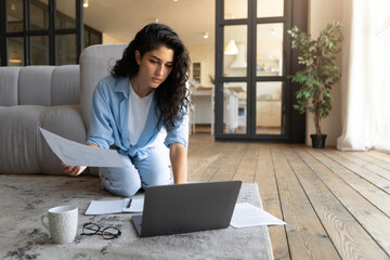 Serious young Cauasian woman working or studying on laptop from home, looking through documents,...