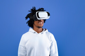 Cool African American teen guy wearing VR headset, using virtual reality for entertainment on blue...