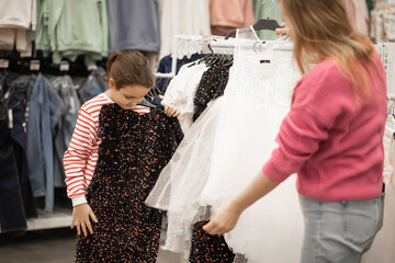 A mother and her young daughter shopping together choose sequined dresses on hangers in a brightly...