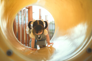 Happy cute Asian toddler girl playing and crawling through a yellow tunnel slide at a sunny summer...