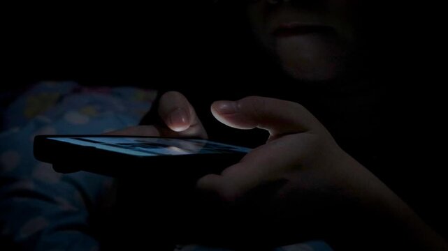Close-up of a woman using a smartphone in a dark bedroom, browsing the internet and scrolling the screen with her finger. Close-up of a female hand scrolling on a mobile phone in a dimly lit room. 
