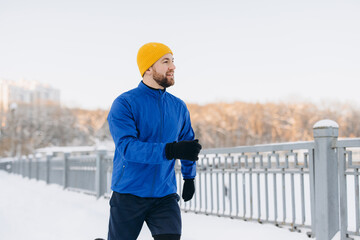 Young man jogging in winter on a snow covered urban path, wearing blue jacket and yellow beanie, smiling and fit