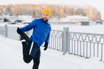 Man in winter sportswear stretching quadriceps on a snowy day, preparing for outdoor exercise and healthy lifestyle