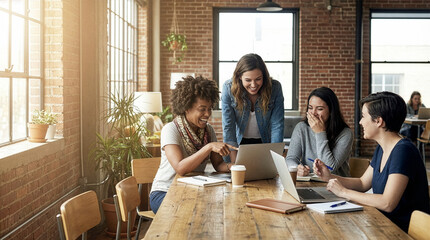 Candid shot of four happy diverse women laughing and pointing at a laptop screen during a creative meeting in a rustic industrial office with brick walls and large windows