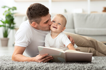 Naklejka premium Child Care. Loving Young Father Reading Book And Kissing His Cute Toddler Son While Relaxing At Home Together, Millennial Dad Spending Time With Infant Baby, Enjoying Paternity Leave, Closeup