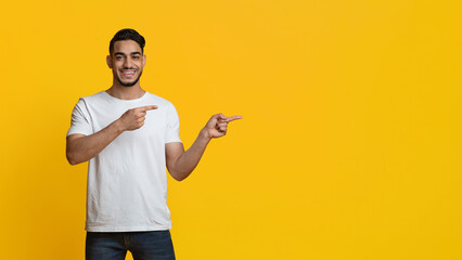 Cheerful middle eastern millennial guy in casual outfit pointing at copy space over yellow studio...