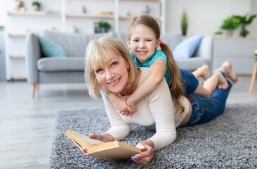Naklejka premium Story Time. Portrait of excited mature woman and cute little girl reading book together lying on rug floor carpet in living room, looking at camera, staying at home, free copy space