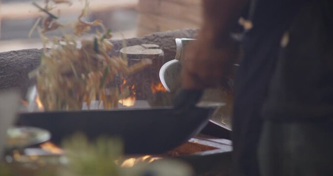 A vendor cooks pad thai noodles in a hot wok over open flame at a local market in Thailand, showing intense heat, metal pans, and traditional street food preparation.