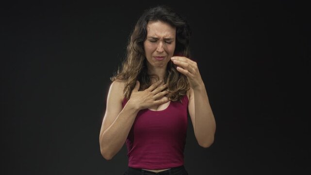 Young woman holds hand to chest with wincing expression for heartattack in black studio setting; pain.