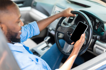Naklejka premium Portrait of happy African American male driver holding and using smartphone with black blank screen, mock up. Closeup of cellphone with empty display, guy using mobile app for navigation, copy space