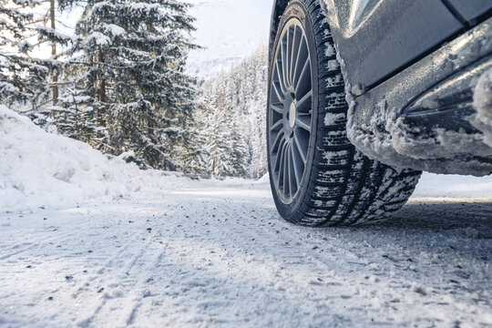 Close-up of a winter tire on a grey metallic car with alloy sport wheels in frog perspective on a snow-covered mountain forest road with snow-laden trees in the background