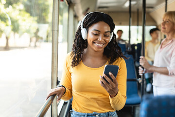Portrait of happy smiling black female passenger traveling in a train or bus, listening to music in wireless headset and using mobile phone, sending sms, standing in public transport near window