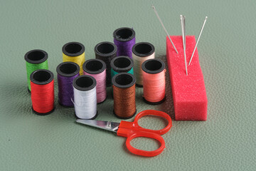 Spools of thread, scissors and red foam needle stand on green leather background.