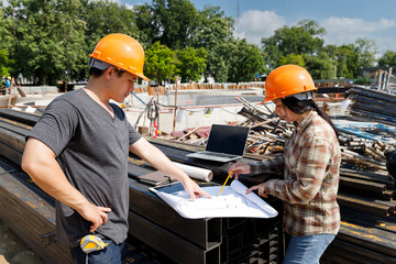Asian construction engineers discussing project blueprint on steel beams with laptop at site. Two civil architects checking building plans on metal stack at industrial construction work.