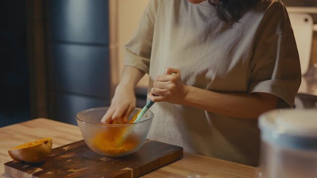 Medium shot of woman grating fresh butternut squash in a home kitchen. Half of the squash lies on wooden cutting board. Blurred domestic background, warm atmosphere, healthy organic meal preparation.