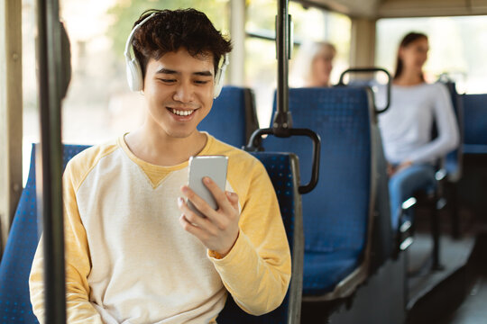 Portrait of happy smiling Asian male passenger traveling in a train or bus, listening to music in wireless headset and using mobile phone, sending sms, sitting in public transport seat, enjoying ride