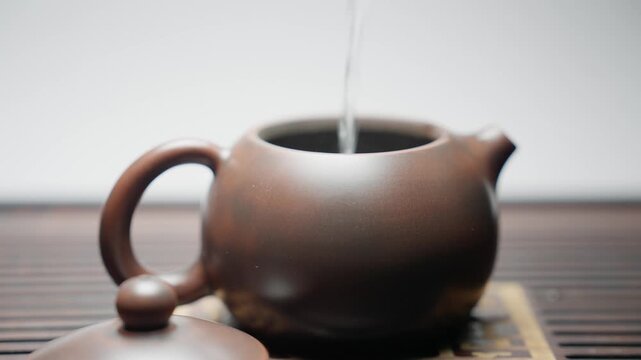 Close-up side view in slow motion. Boiling water is poured into a brown clay teapot on a traditional tea board, followed by placing the lid. Captures a key step of tea brewing.