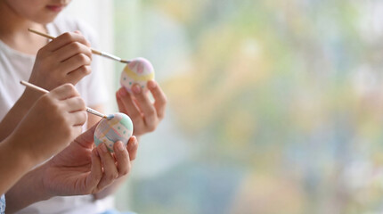 Kids paint Easter eggs with brushes. Pastel egg designs in hands, egg craft for Easter family activity, spring DIY background with copy space, soft window light, joyful moment, cozy concept